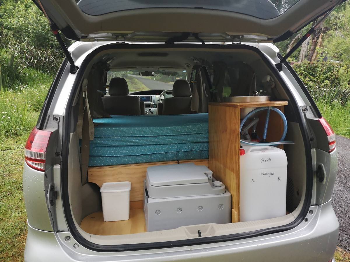 Tailgate open on the JNQ545 campervan parked on a grass verge, kitchen unit and water container inside