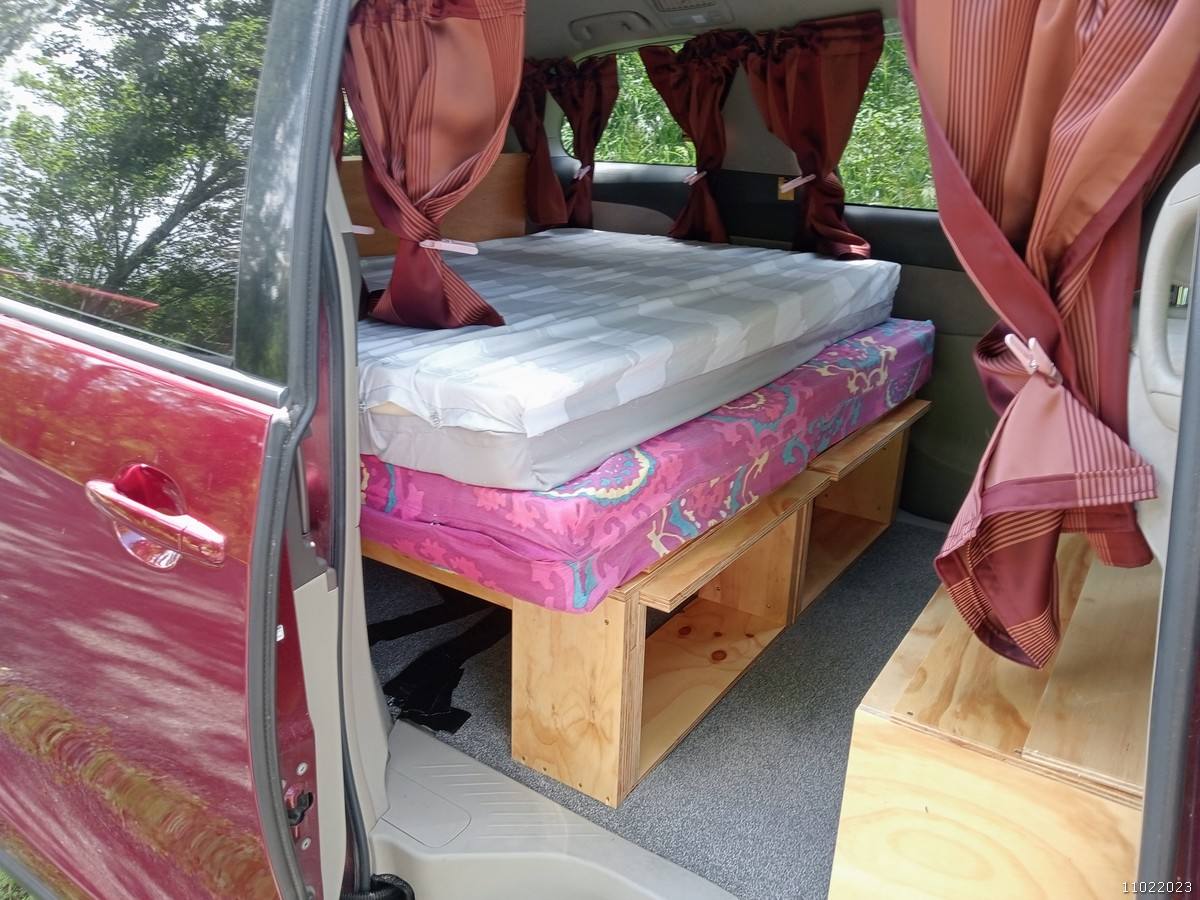 Side door open on the JAK653 Toyota Estima campervan, bed made up with red curtains tied back