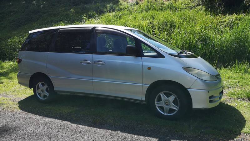 Silver 2000 Toyota Estima self-contained campervan for sale, side view parked on grass below a bush bank