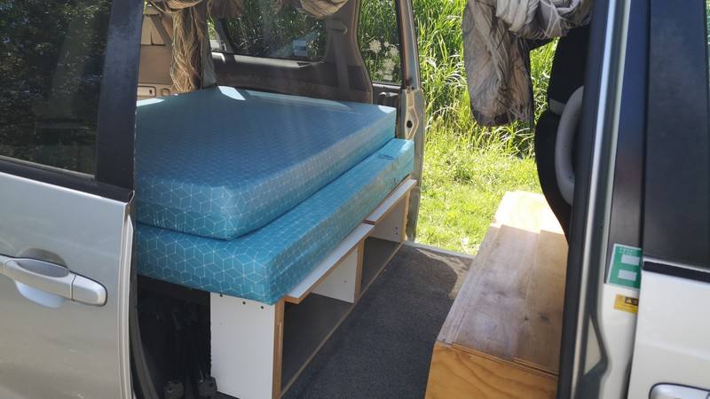 Side door open on the silver Toyota Estima campervan, bed made up over the storage shelves, step board alongside