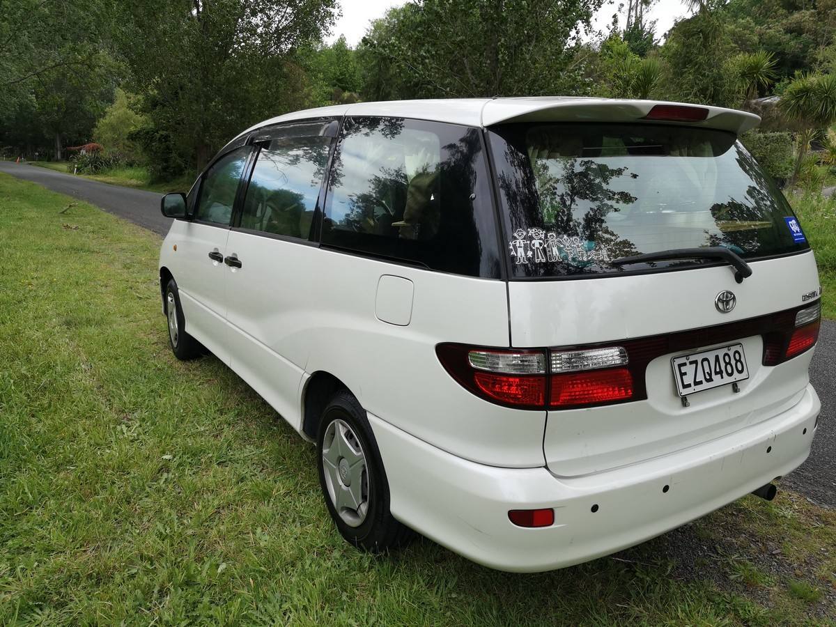 Rear three-quarter view of the white Toyota Estima campervan EZQ488 on a tree-lined road