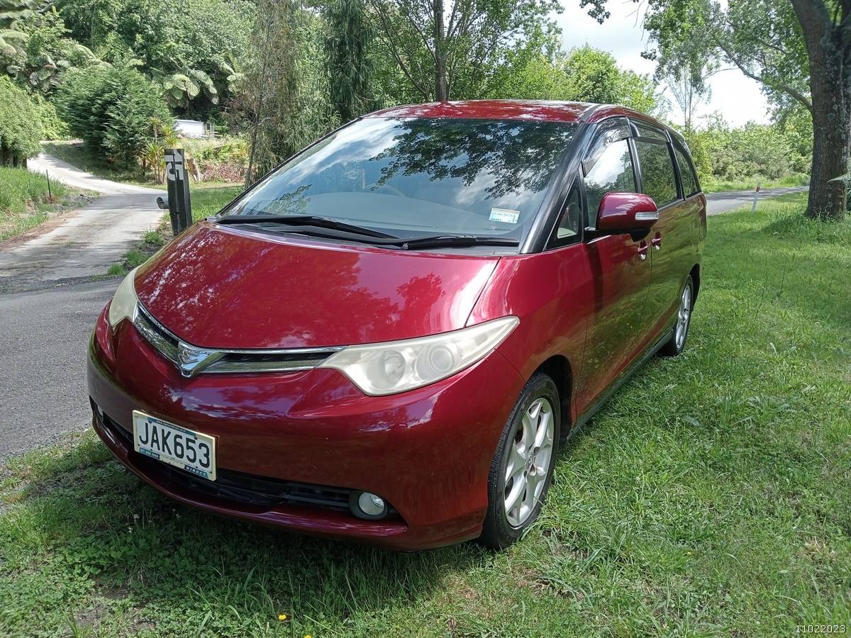 Front three-quarter view of the red JAK653 Toyota Estima campervan on a tree-lined drive