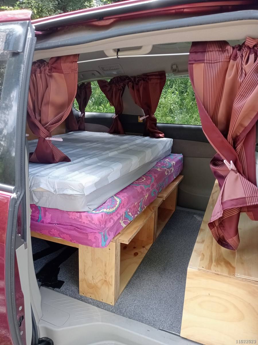 Side door open on the red JAK653 campervan, double bed on its plywood base with storage underneath