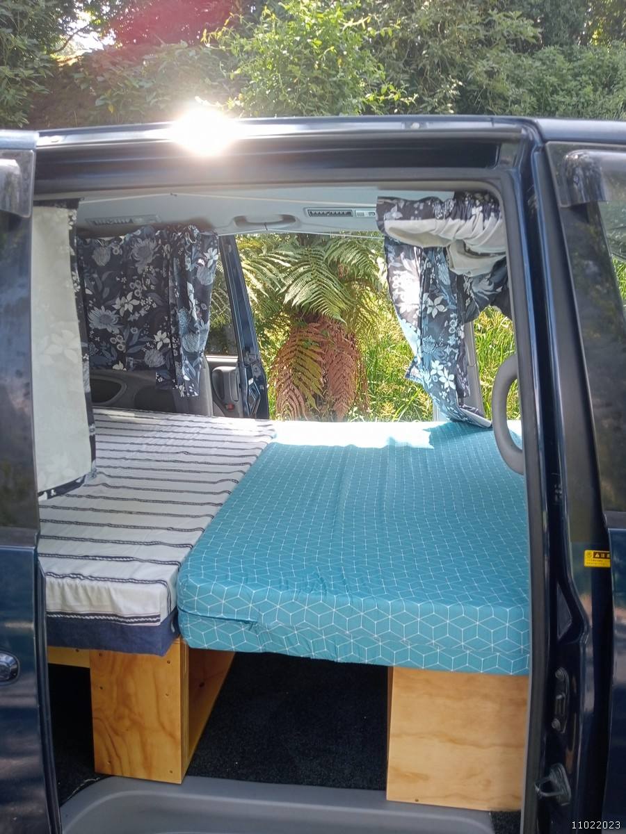 Side door open on the dark blue FDK492 campervan, double bed made up with the ferns behind