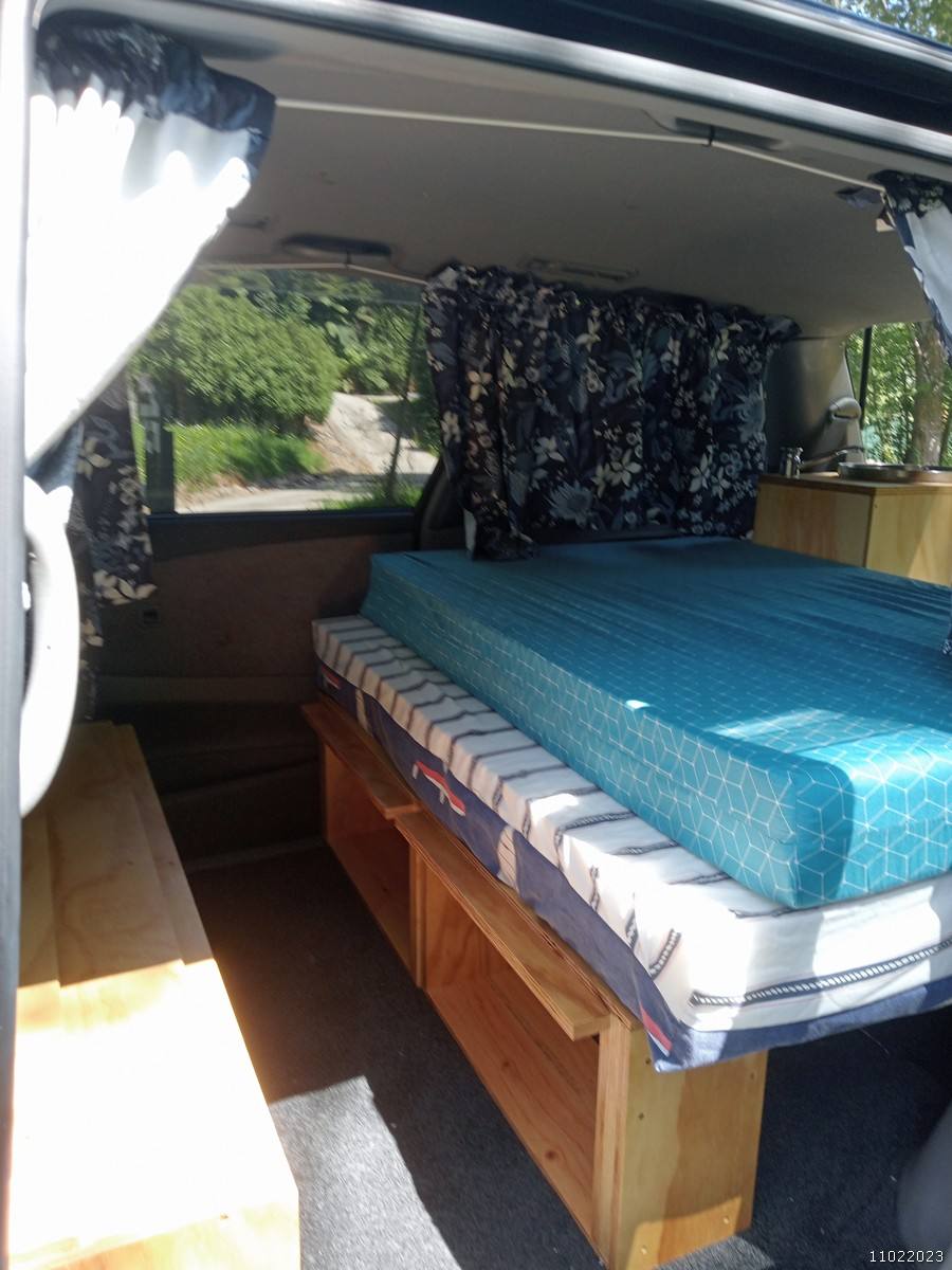 Inside the FDK492 campervan, bed made up on its plywood base with floral curtains at the windows